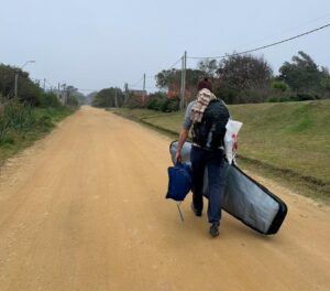 man carrying a surfboard and gear on a dirt road.